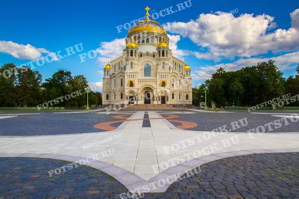 Фотография представляет собой снимок Никольского морского собора в Кронштадте, одной из знаковых достопримечательностей Санкт-Петербурга. Собор выделяется своими светлыми стенами и золотыми куполами, которые великолепно смотрятся на фоне синего неба с белыми облаками. Вид собора открыт, что позволяет оценить всю его архитектурную красоту, а мощеная площадь перед собором придает композиции чувство пространства и порядка. Это изображение идеально подходит для путеводителей, религиозных и культурных изданий, а также может использоваться для украшения интерьеров или в качестве образовательного материала.